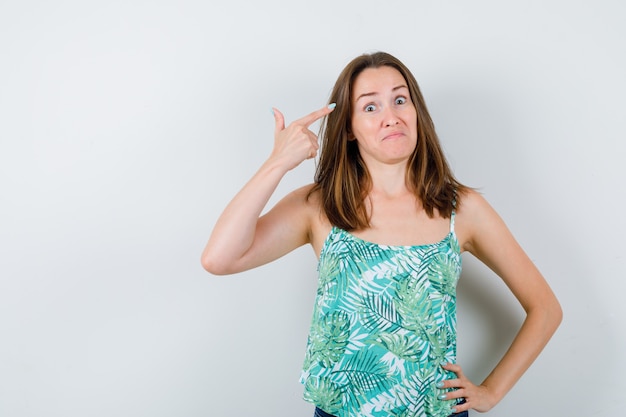 Portret van een jonge dame die naar het hoofd wijst terwijl ze de hand op de heup in de blouse houdt en besluiteloos vooraanzicht kijkt