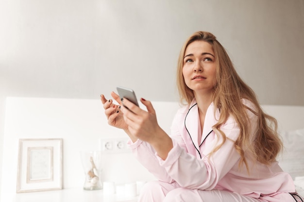 Portret van een jonge dame die op bed zit met een mobiele telefoon in handen en thuis zorgvuldig opzij kijkt