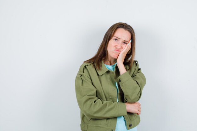 Portret van een jonge dame die op haar wang leunt in t-shirt, jas en er somber vooraanzicht uitziet