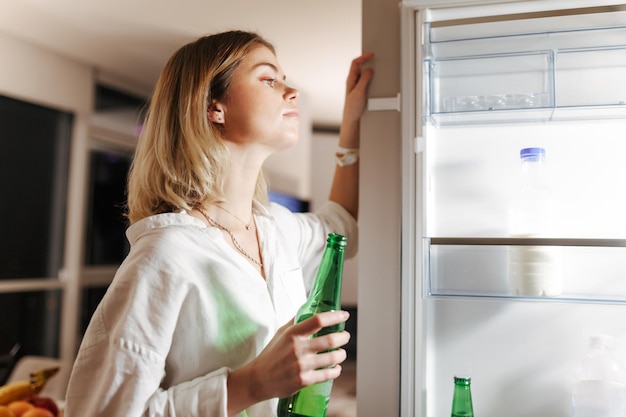 Portret van een jonge dame die 's nachts in de keuken staat en dromerig in de open koelkast kijkt terwijl ze thuis bier in de hand houdt