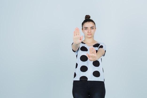 Portret van een jonge dame die stopgebaar in t-shirt, jeans toont en ernstig vooraanzicht kijkt