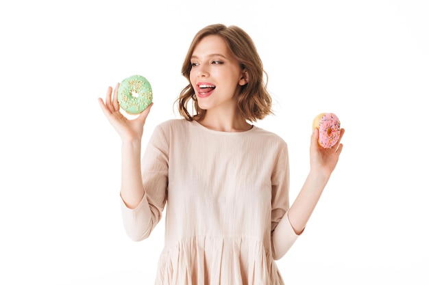 Portret van een jonge dame in een roze jurk die staat en dromerig op donuts kijkt op een witte achtergrond geïsoleerd