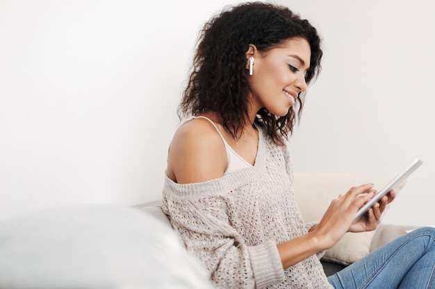 Portret van een jonge dame met donker krullend haar in oortelefoons zittend op de bank met tablet in handen thuis