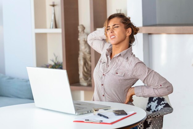 Portret van een jonge gestresste vrouw die aan een thuisbureau voor een laptop zit en een pijnlijke rug aanraakt met een gepijnigde uitdrukking die lijdt aan rugpijn na het werken op een laptop