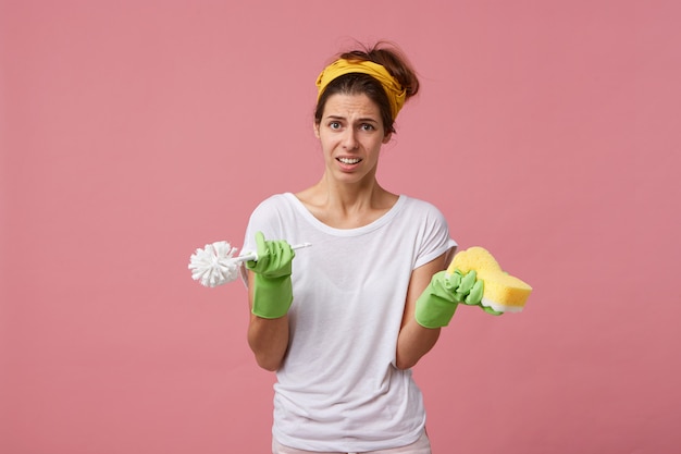 Portret van een jonge huisvrouw die een wit T-shirt, groene handschoenen en een gele sjaal op het hoofd draagt met borstel en spons met fronsend gezicht dat niet wil schoonmaken