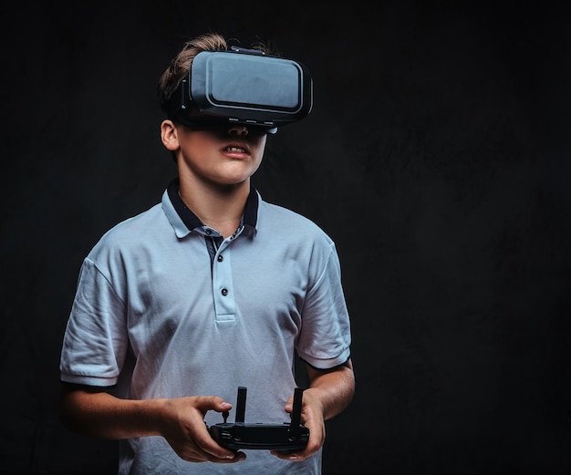 Portret van een jonge jongen gekleed in een wit t-shirt met een virtual reality-bril en een afstandsbediening vast. Geïsoleerd op de donkere achtergrond.