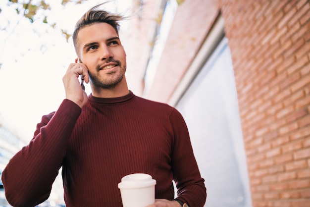 Portret van een jonge knappe man praten aan de telefoon terwijl hij een kopje koffie buiten op straat houdt. Communicatie concept.