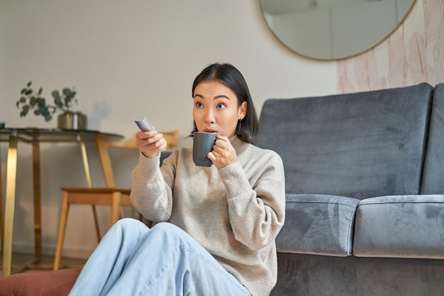 Gratis foto portret van een jonge koreaanse vrouw die televisie kijkt met afstandsbediening en verbaasd kijkt naar tv-scherm sp