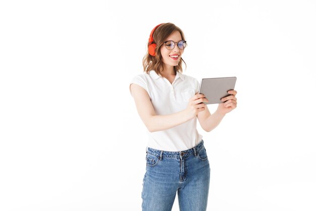 Portret van een jonge lachende dame in brillen en rode koptelefoons die staan en gelukkig kijken in tablet op een witte achtergrond geïsoleerd