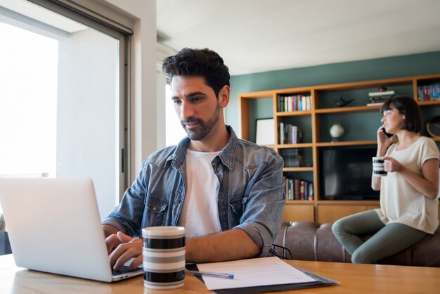 Portret van een jonge man aan het werk met een laptop vanuit huis terwijl vrouw praten over de telefoon op de achtergrond