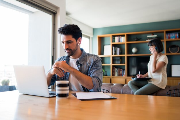 Portret van een jonge man aan het werk met een laptop vanuit huis terwijl vrouw praten over de telefoon