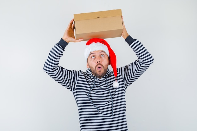 Gratis foto portret van een jonge man die een kartonnen doos op zijn hoofd houdt in een hoodie, een kerstmuts en zich afvraagt