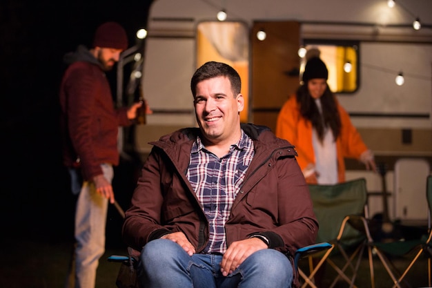 Portret van een jonge man die lacht zittend op een campingstoel met een vriend met een biertje op de achtergrond. Retro camper.