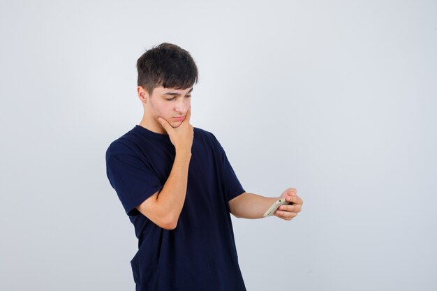 Portret van een jonge man die mobiele telefoon bekijkt, hand op kin in zwart t-shirt houdt en doordacht vooraanzicht kijkt