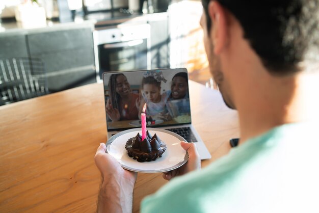 Portret van een jonge man die verjaardag viert op een videogesprek vanuit huis met laptop en een taart. Nieuw normaal levensstijlconcept.