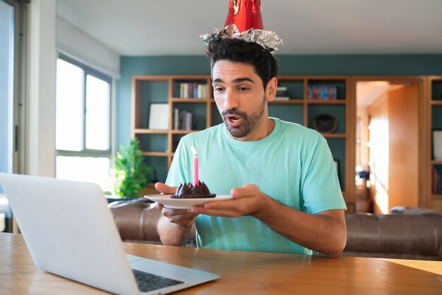 Portret van een jonge man die verjaardag viert tijdens een videogesprek vanuit huis met laptop en een taart. Nieuw normaal levensstijlconcept.
