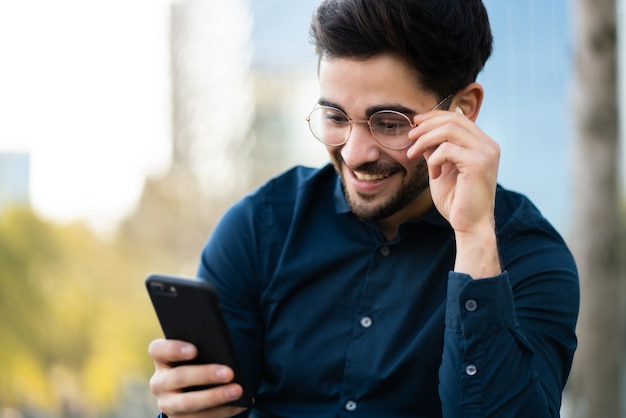 Portret van een jonge man met zijn mobiele telefoon zittend op de bank buiten. Stedelijk concept.