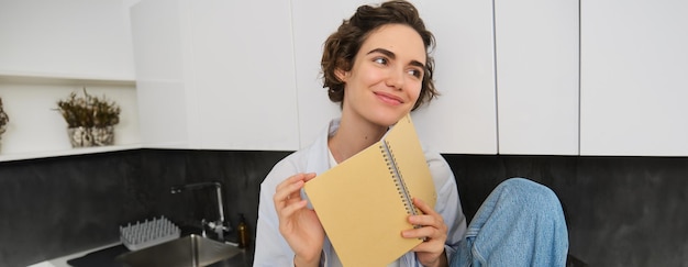 Gratis foto portret van een jonge moderne vrouw die in de keuken zit en bladert en glimlachend een dagboek leest