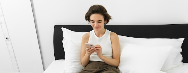 Portret van een jonge mooie brunette vrouw die op bed zit met een smartphone met behulp van een mobiele telefoonapp