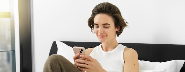 Portret van een jonge mooie brunette vrouw die op bed zit met een smartphone met behulp van een mobiele telefoonapp