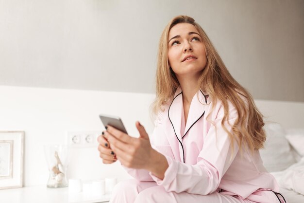 Portret van een jonge, mooie dame die op bed zit met een mobiele telefoon in handen en thuis dromerig opzij kijkt