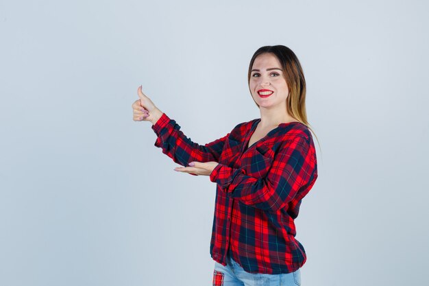 Portret van een jonge, mooie vrouw die de palm opzij spreidt, de duim in een casual shirt laat zien en er vrolijk vooraanzicht uitziet