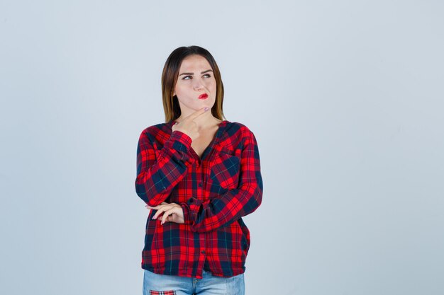 Portret van een jonge, mooie vrouw die de vinger op de kin houdt in een casual shirt en er doordacht vooraanzicht uitziet