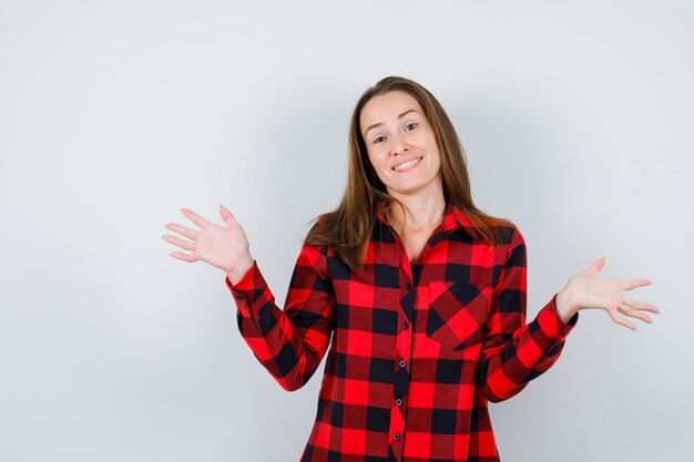 Portret van een jonge, mooie vrouw die een hulpeloos gebaar in een casual shirt toont en er vrolijk vooraanzicht uitziet