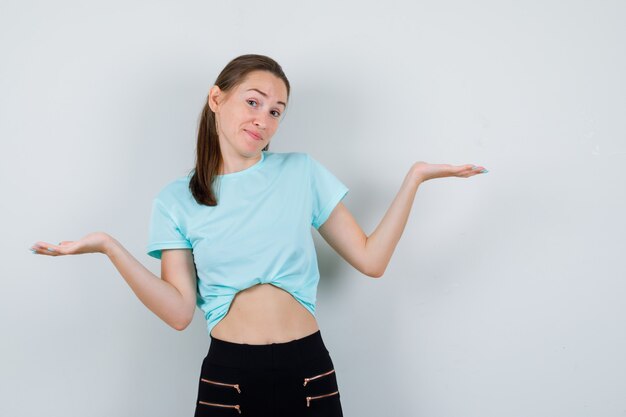 Portret van een jonge, mooie vrouw die een hulpeloos gebaar toont in een t-shirt, broek en een vrolijk vooraanzicht