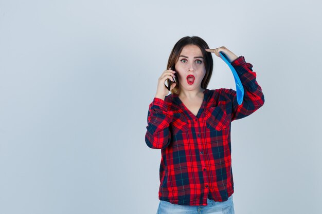 Portret van een jonge, mooie vrouw die een map vasthoudt terwijl ze aan het telefoneren is, de vinger op het hoofd houdt in een casual shirt, jeans en een geschokt vooraanzicht