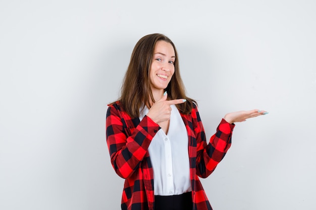 Portret van een jonge, mooie vrouw die naar rechts wijst, de handpalm opzij spreidt in een casual outfit en er vrolijk vooraanzicht uitziet