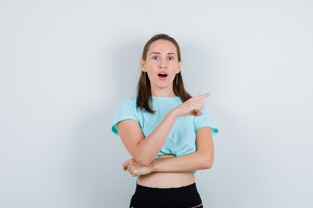 Portret van een jonge, mooie vrouw die recht in een t-shirt wijst en verbijsterd vooraanzicht kijkt