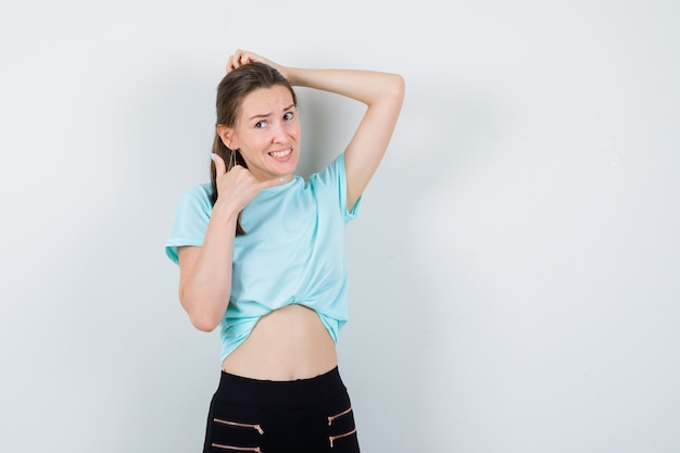 Portret van een jonge, mooie vrouw met de hand op het hoofd, met telefoongebaar in t-shirt, broek en boos vooraanzicht