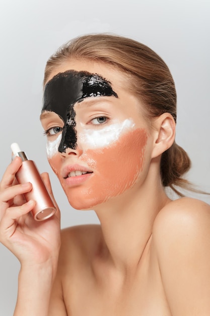 Portret van een jonge mooie vrouw met verschillende cosmetische maskers op het gezicht die schoonheidsvloeistof in de hand houdt terwijl ze dromerig in de camera kijkt over een grijze achtergrond