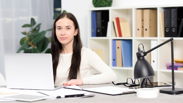 Portret van een jonge onderneemster die met laptop op bureau aan camera kijkt