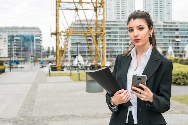 Portret van een jonge onderneemster die slimme telefoon en in hand omslag houdt