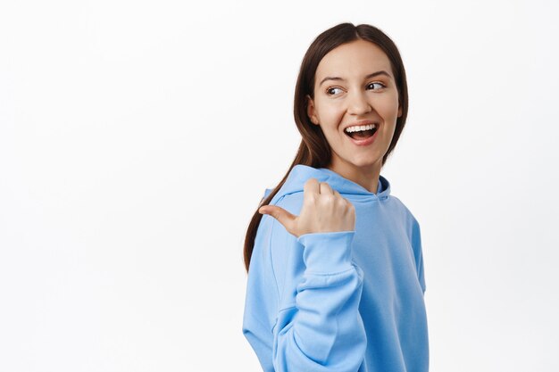 Portret van een jonge openhartige vrouw, gelukkig glimlachend, naar links wijzend en achter haar schouder kijkend naar de verkoopbanner, staande in hoodie tegen de witte muur.