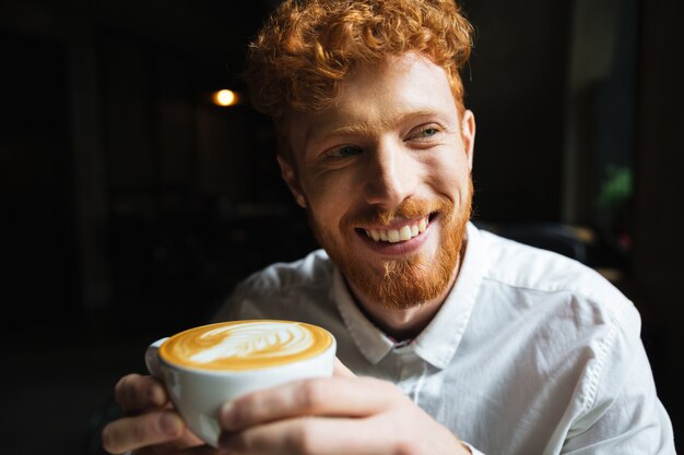 Portret van een jonge redhead bebaarde man met charmante glimlach in wit overhemd met koffiekopje, opzij kijken
