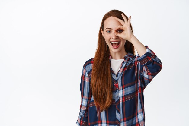 Portret van een jonge roodharige vrouw die een goed teken nul gebaar toont en ja zegt, kijk verbaasd en gelukkig in een geruit overhemd tegen een witte achtergrond