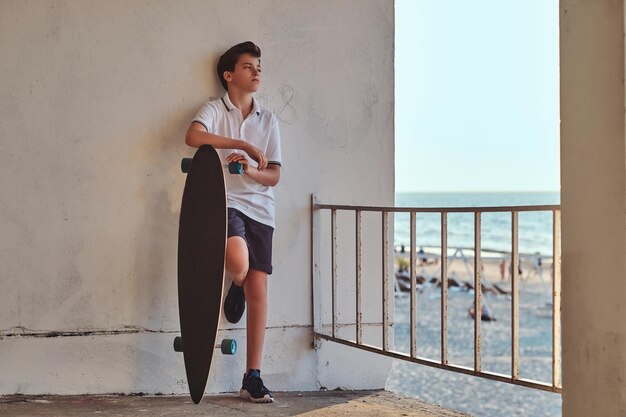 Portret van een jonge skaterjongen gekleed in een t-shirt en korte broek, leunend op een muur en houdt een skateboard vast tegen de achtergrond van een zeekust.