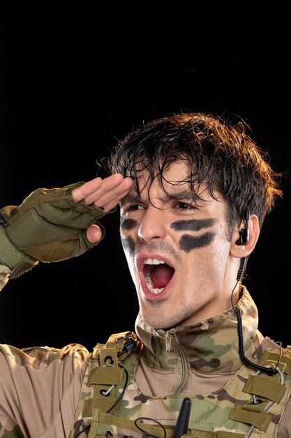 Portret van een jonge soldaat in camouflage die op zwarte muur salueert