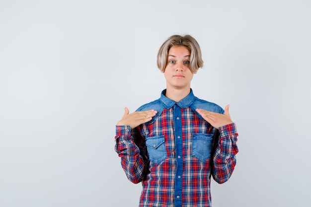 Portret van een jonge tienerjongen die naar zichzelf wijst in een geruit overhemd en een verbaasd vooraanzicht kijkt