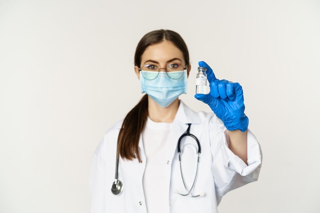 Portret van een jonge vrouw, arts met medisch gezichtsmasker en uniform, met vaccin, covid-19 vaccinatiecampagne, staande op een witte achtergrond.