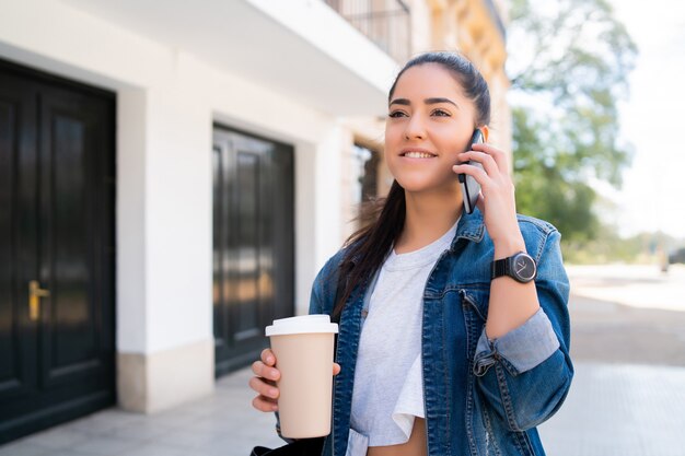 Portret van een jonge vrouw die aan de telefoon praat en een kopje koffie vasthoudt terwijl ze buiten op straat staat