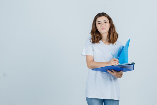 Portret van een jonge vrouw die aantekeningen maakt over de map in wit t-shirt, spijkerbroek en op zoek vrolijk vooraanzicht