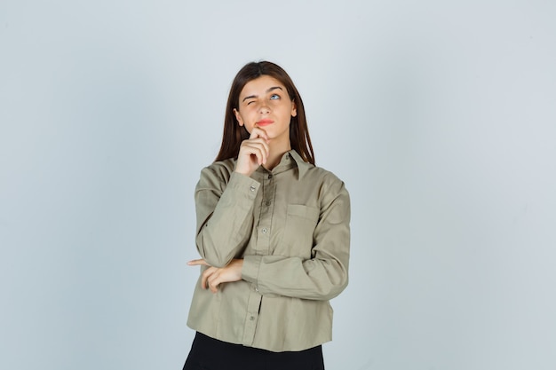 Portret van een jonge vrouw die de hand op de kin in shirt, rok houdt en er attent vooraanzicht uitziet