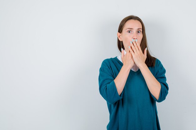 Portret van een jonge vrouw die de handen op de mond houdt in een trui over een wit overhemd en er verbaasd uitziet aan de voorkant