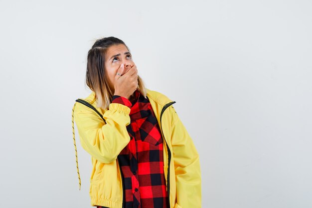 Portret van een jonge vrouw die de mond bedekt met handen, omhoog kijkt in een geruit hemd, een jas en een geschokt vooraanzicht