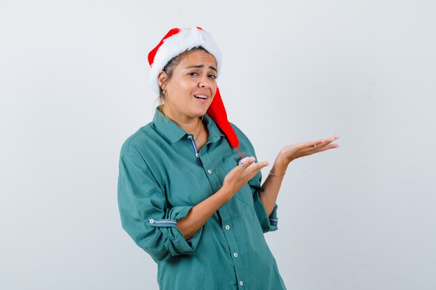 Portret van een jonge vrouw die doet alsof ze iets in shirt, kerstmuts laat zien en er verbaasd uitziet