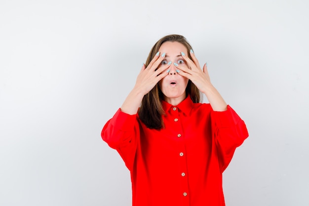 Portret van een jonge vrouw die door de vingers in een rode blouse kijkt en zich afvraagt vooraanzicht